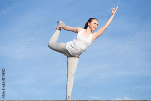 Sporty young woman practicing yoga, standing in Lord of the Dance exercise, Natarajasana pose, working out, wearing sportswear, outdoor, clear blue sky background