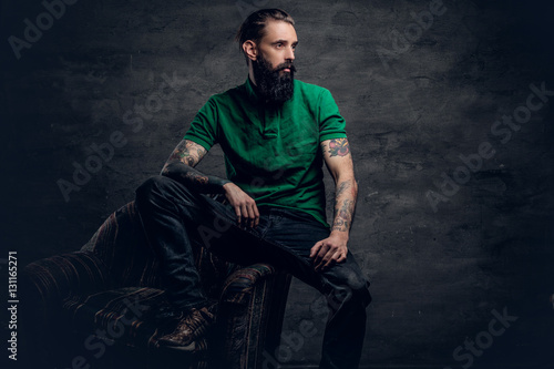 Tattooed bearded male sits on a chair.