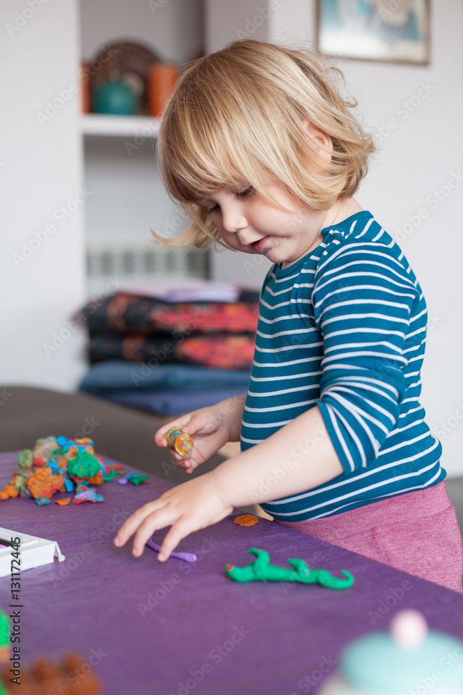 blonde two years old child with striped blue and white sweater inside home playing modeling figures with plasticine on purple table 
