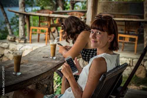 Best friends having fun, laughing. Tropical cafe on the cliff in Nusa Lembongan, Indonesia. Girls drinking ice coffee.