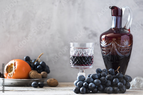 old fashioned crystal glass jug with crystal glass of red wine and and metallic plate with bunch of black grapes, persimmon and nuts on wooden table and cones around with grey wall as a background 