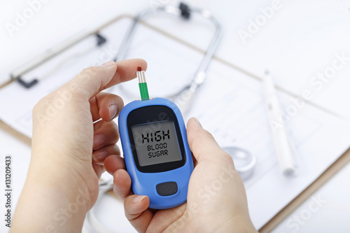 Hand holding a blood glucose meter measuring blood sugar, the background is a stethoscope and chart file