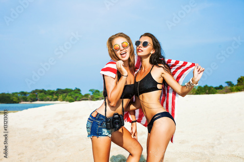 Two cool trendy hipster girls at the beach enjoying vacation on a tropical island, perfect tanned body, healthy skin, sexy stylish casual wear, bikini, blond and brunette with the camera, sunglasses