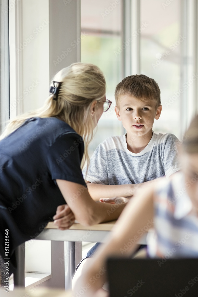 Teacher assisting boy at desk with girl in foreground Teacher assisting boy at desk with girl in foreground