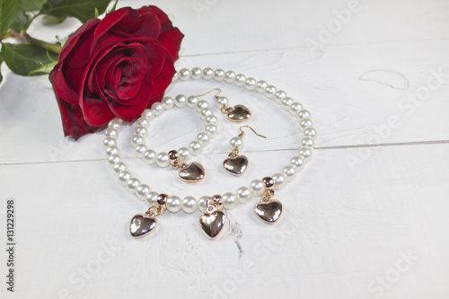 Pearl necklace and earrings with golden hearts and red rose on white wood