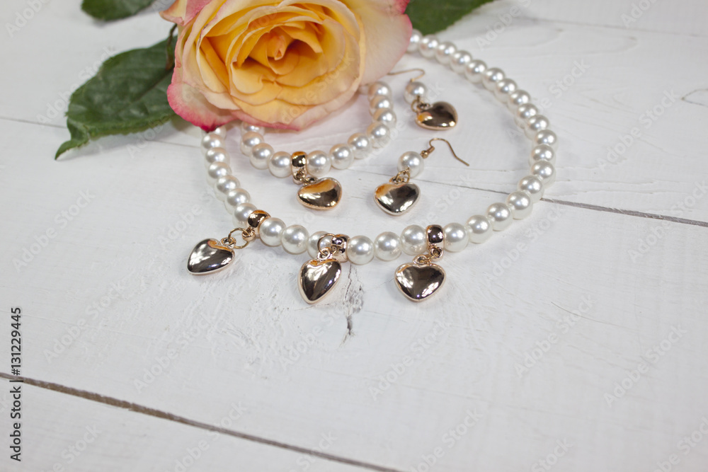 Pearl necklace and earrings with golden hearts and yellow rose on white wood