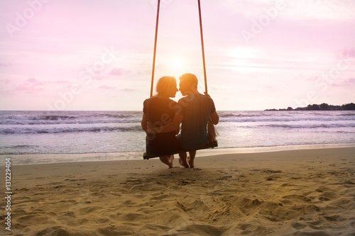 Romantic couple is sitting and kissing on sea beach on rope swing and looking at sunset horizon. Family vacation on honeymoon photo
