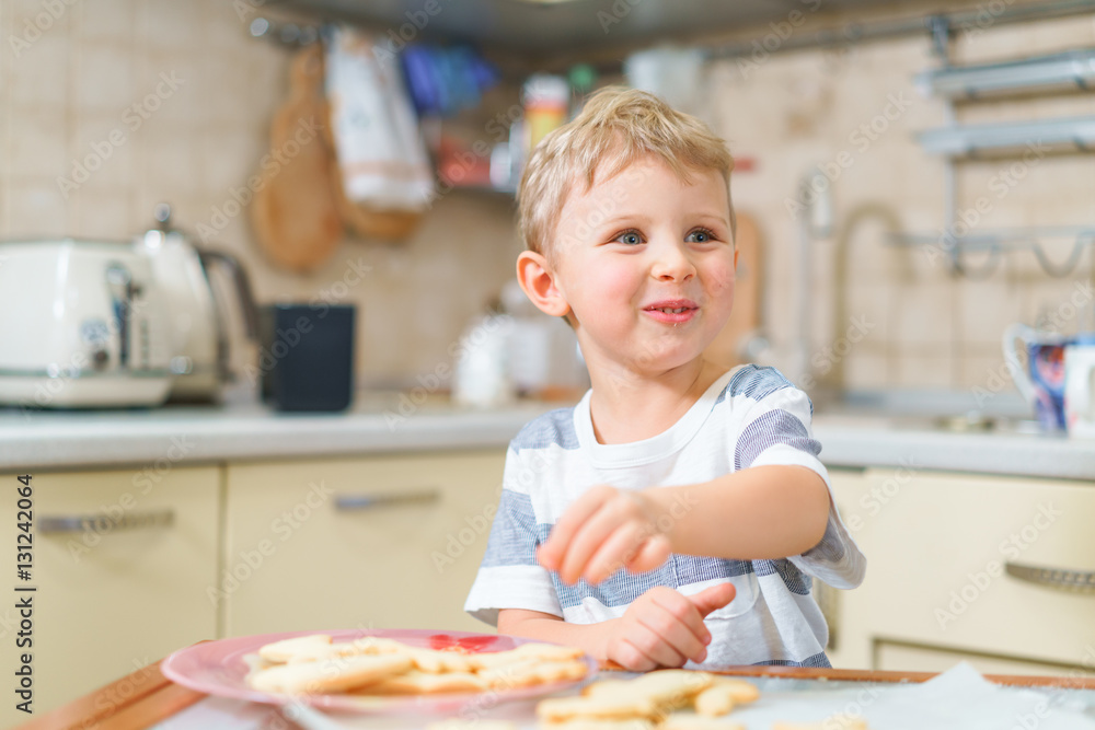 Little blond kid tries freshly baked shortbread, sitting at the kitchen table. Smiling face. Little blond kid tries freshly baked shortbread, sitting at the kitchen table. Smiling face.