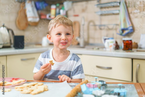 Little blond kid tries freshly baked shortbread, sitting at the kitchen table. Smiling face.