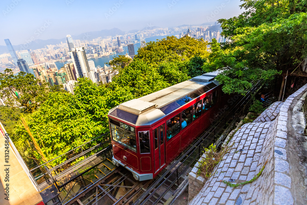 Obraz premium Popularny czerwony tramwaj Peak dociera do końca Victoria Peak, najwyższego szczytu wyspy w Hongkongu, z panoramą miasta w tle. Słoneczny dzień.