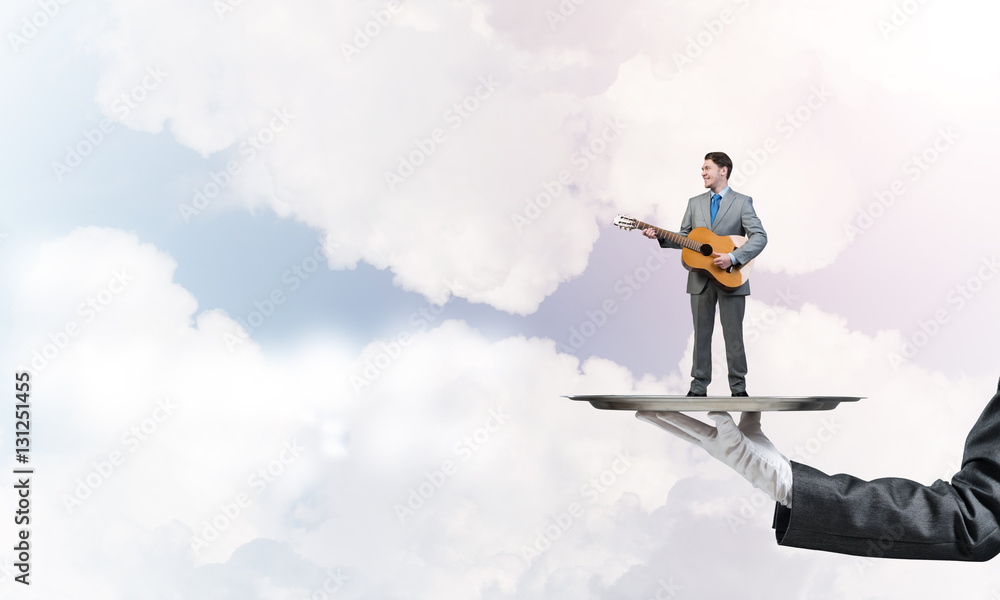 Businessman on metal tray playing acoustic guitar against blue sky background Businessman on metal tray playing acoustic guitar against blue sky background