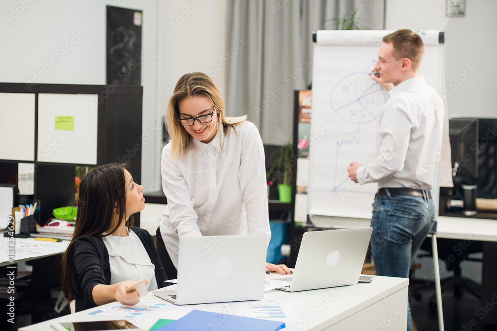 Obraz premium Two beautiful office workers having conversation while man colleague drawing business strategy on flip chart.