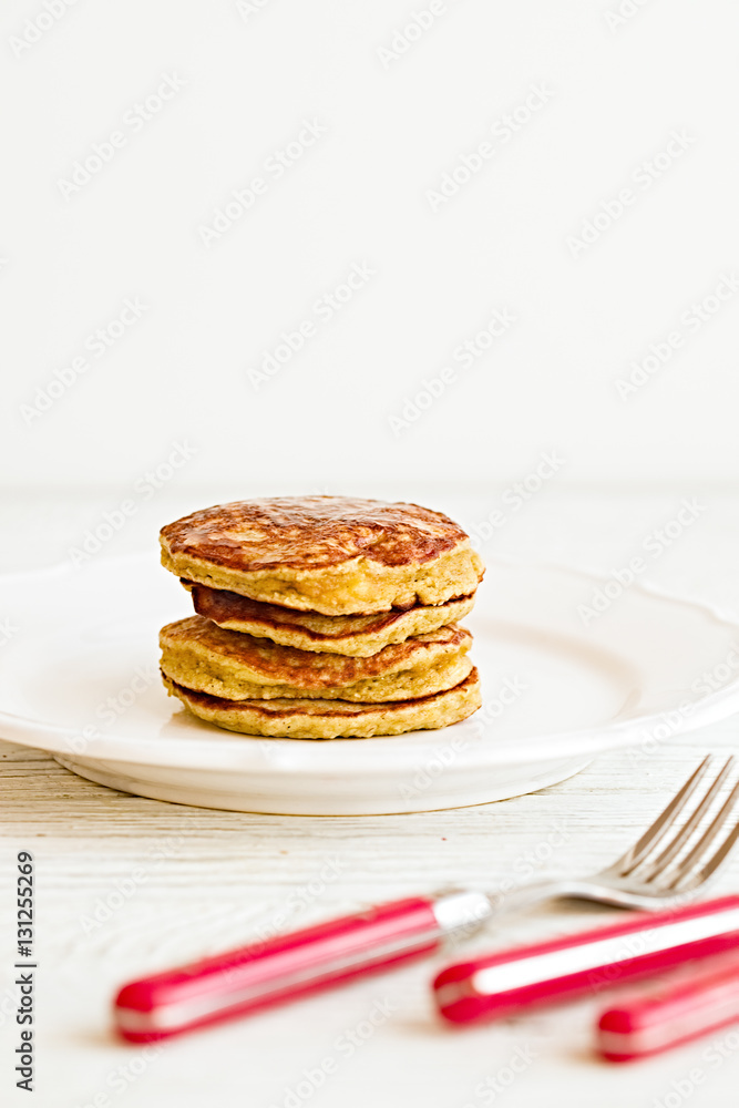 Plain paleo pancakes with banana and coconut on a white plate with red fork and knife. Bright healthy breakfast. White wooden table Plain paleo pancakes with banana and coconut on a white plate with red fork and knife. Bright healthy breakfast. White wooden table