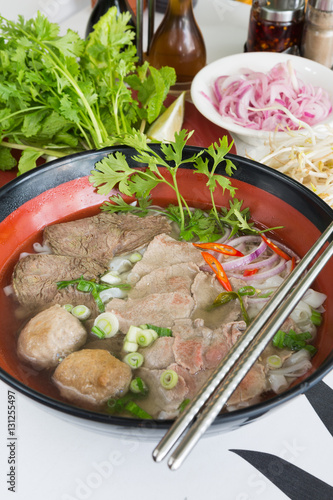 A bowl of traditional vietnamese beef and chicken pho street food, surrounded by fresh herbs. Pho is a vietnamese noodle soup consisting of meat, herbs, chilli, coriander and dumplings.