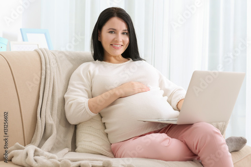 Beautiful young pregnant woman with laptop sitting on sofa at home