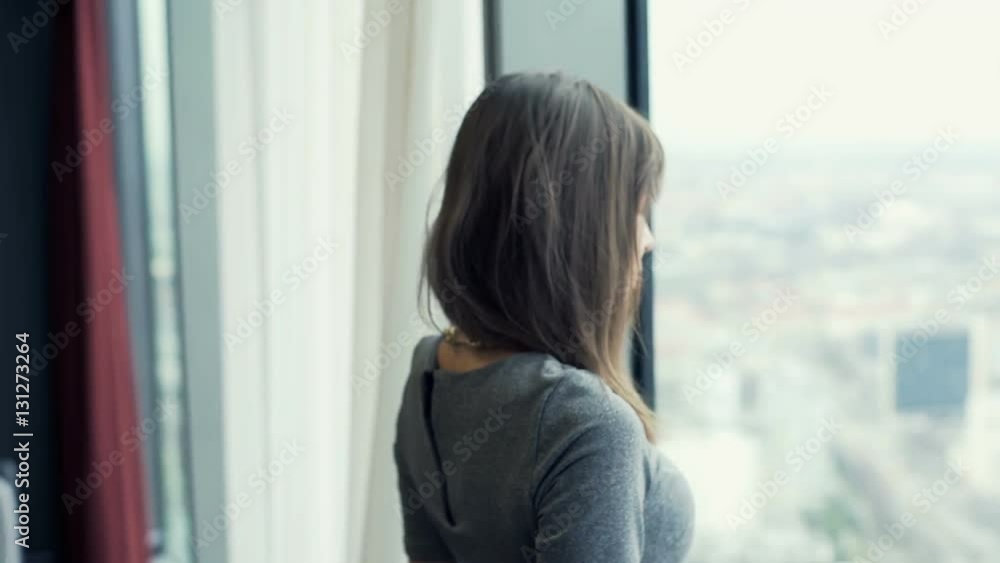 Young woman unveil curtains and look at the city from her apartment
Young woman unveil curtains and look at the city from her apartment