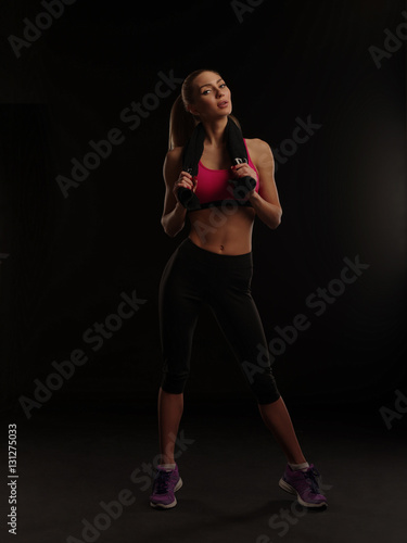 Beautiful athletic young woman with slim gorgeous body and collected hair wearing the sportswear is posing with a towel on the dark black background, fitness and workout