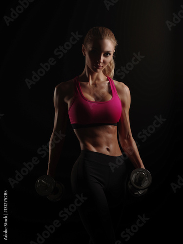 Beautiful muscular young woman with slim fitness body wearing the sportswear is doing exercises with dumbbells on the dark black background in the studio, sweat on the abdominal, sports equipment