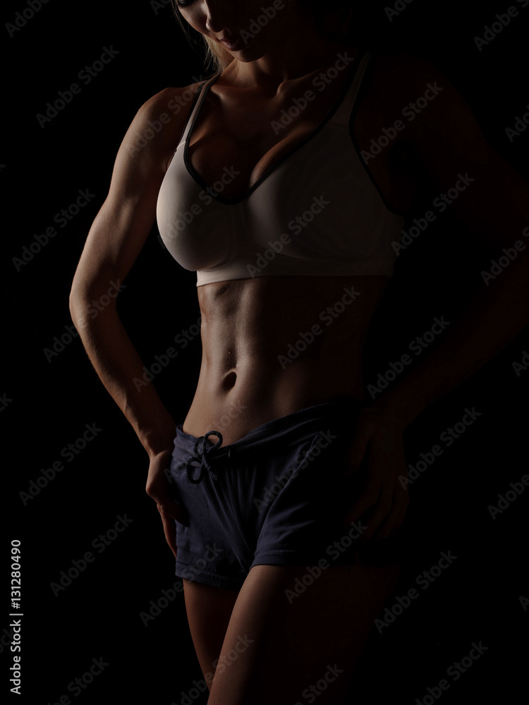 Young athletic and sexy woman in a white sports shirt and blue shorts in the studio on a black background shows strong biceps and a flat belly with the perfect abdominal muscles, dark black background