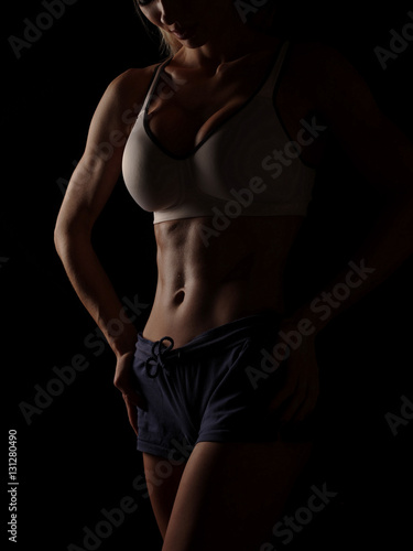 Young athletic and sexy woman in a white sports shirt and blue shorts in the studio on a black background shows strong biceps and a flat belly with the perfect abdominal muscles, dark black background