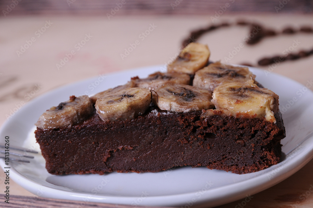 A piece of chocolate cake with banana on a white saucer. Close-up. Heart of coffee beans at the background on wooden tray