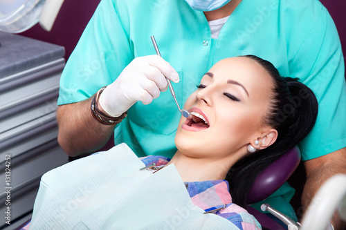 Male dentist examines the mouth of the young beautiful woman patient with perfect straight white teeth on the dentist's chair with the help of dental mirror. Healthcare, medicine. photo