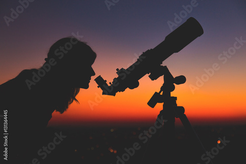 Girl looking at the stars with telescope beside her and de-focused city lights. 