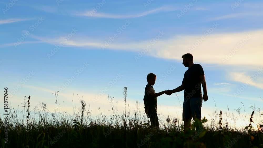 Anonymous father and son shakes hands in agreement. Happy family having fun in evening beautiful nature landscape. Silhouettes of man and boy over sunset blue sky background. Real time video footage. Anonymous father and son shakes hands in agreement. Happy family having fun in evening beautiful nature landscape. Silhouettes of man and boy over sunset blue sky background. Real time video footage.