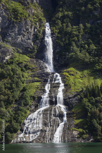 Beautiful waterfall from small cleft to green lake with green trees and picturesque rocks around  Norway