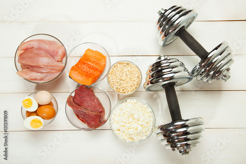 Protein diet, fish, cheese, eggs, meat, chicken and dumbbells on a white wooden background