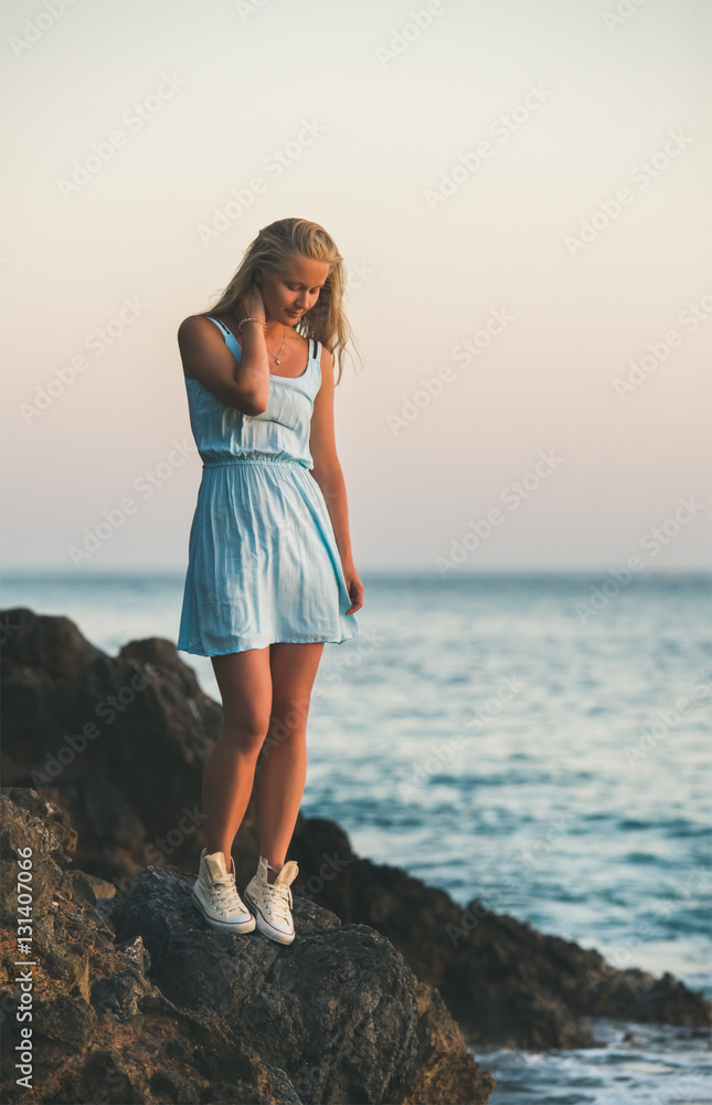 Young blond tourist woman in blue dress standing on natural rocks by the sea at sunset and looking down. Kleopatra beach, Alanya, Mediterranean region, Turkey
