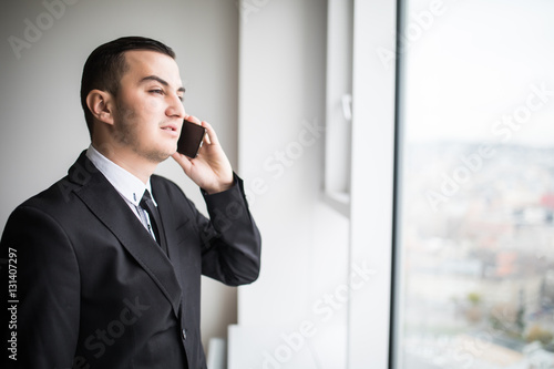 Handsome young businessman standing next to the large windows of his top floor office, looking at the view of the city while talking on his mobile phone