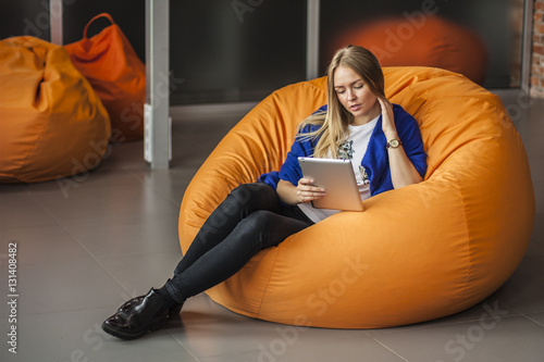 Armchair traditional bean bag for fun and relaxation  orange color. Young woman  using tablet screen while sitting on big cushioned frameless chair. 