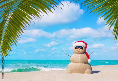 Sandy Snowman in Christmas Santa hat and sunglasses at ocean tropical palm beach
