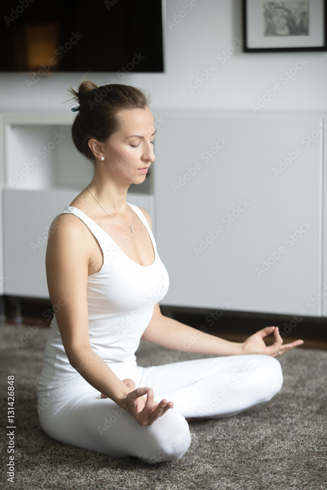 Sporty attractive young woman practicing yoga, sitting in Lotus exercise, Padmasana pose, working out, wearing white sportswear, indoor full length, home interior background Sporty attractive young woman practicing yoga, sitting in Lotus exercise, Padmasana pose, working out, wearing white sportswear, indoor full length, home interior background