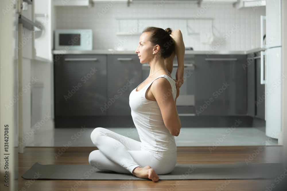 Sporty young attractive woman practicing yoga, sitting in Cow Face exercise, Gomukasana pose, working out, wearing white sportswear, indoor full length, home interior background Sporty young attractive woman practicing yoga, sitting in Cow Face exercise, Gomukasana pose, working out, wearing white sportswear, indoor full length, home interior background