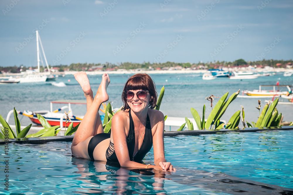 Portrait of beautiful tanned woman in black swimwear relaxing in swimming pool spa. Hot summer day and bright sunny light. Beautiful ocean, sky and asian plants on the background. Tropical island Bali