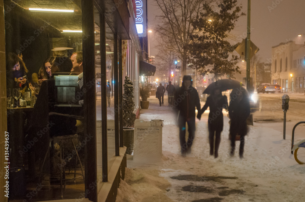 Customers in a bar are seen through a window while pedestrians walk through the snow outdoors.