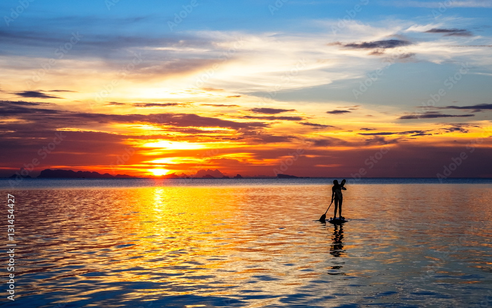 Silhouette of a young woman paddleboarding at sunset with cloudy landscape on the background