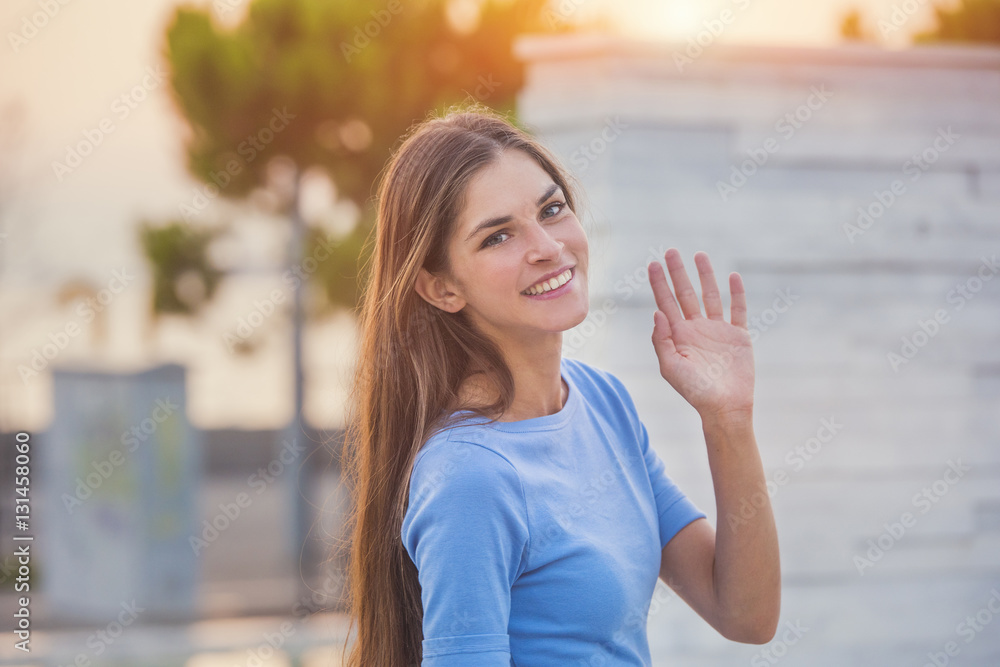 candid portrait of a beautiful young woman waving her hand and smiling, while on a sunset background