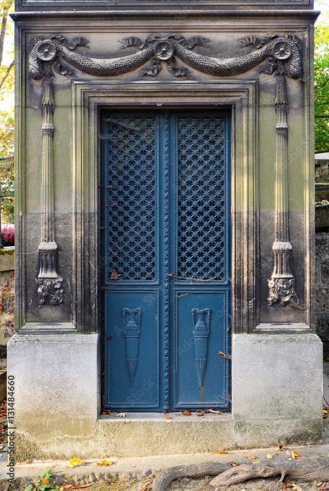 Naklejka premium Rectangular blue double door made of steel. Ornaments of vases, floral and cross pattern. The ornate door frame contains chiseled relief of torches carved in stone. An old tomb / crypt at a graveyard.