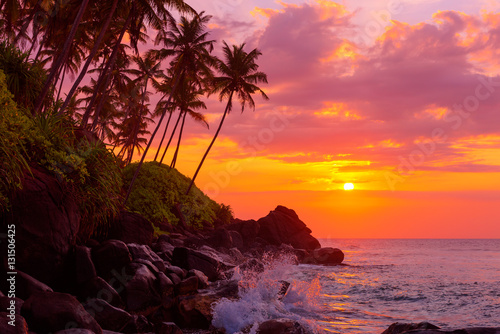 Fototapeta Naklejka Na Ścianę i Meble -  Sunset on tropical coast with rocks in wavy ocean and palm trees on a hill