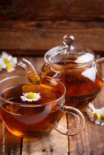 Glass of chamomile tea with full hot teapot and fresh chamomile flowers on vintage old wooden table background
