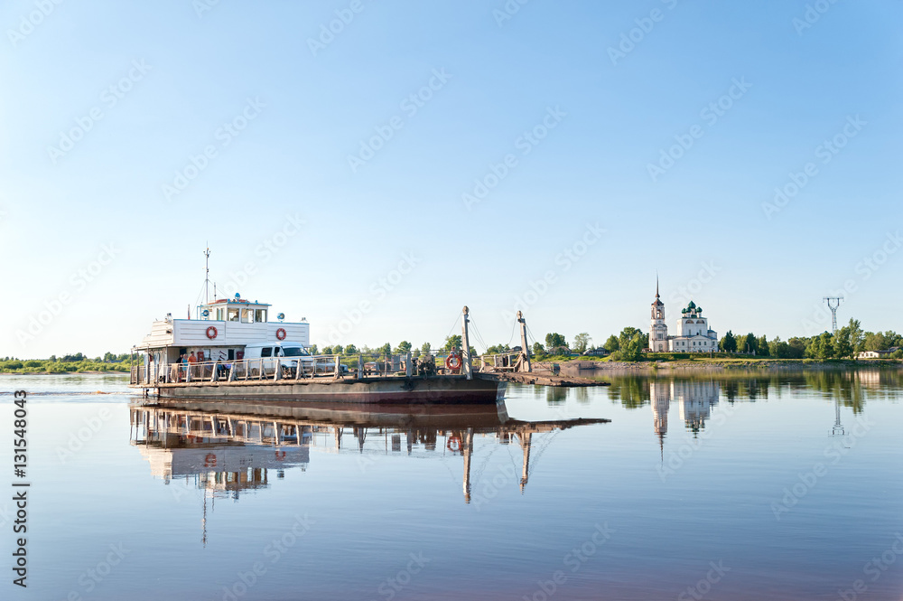 Ferryboat with people and cars goes on Vychegda River with ancient cathedral on riverside in background. Solvychegodsk, Arkhangelsky region, Russia.