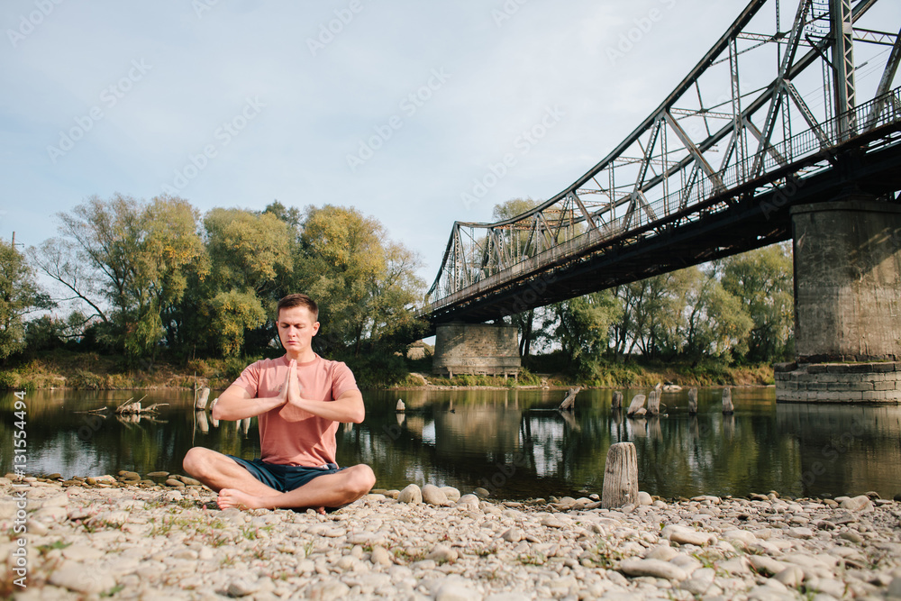 man practices yoga on the river bank near the old bridge
