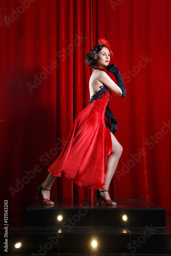 The charming brunette woman in a red dress dancing on the stage on the background of burgundy drapes. © ksi