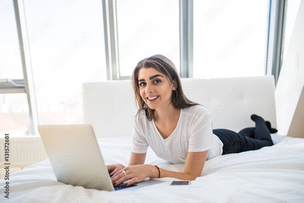 Naklejka premium Portrait of beautiful happy young woman lying in bed and using laptop at home