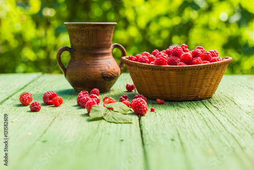 Fresh ripes raspberry in a wcker basket and ceramic jug on a rustic wooden table outdoors.