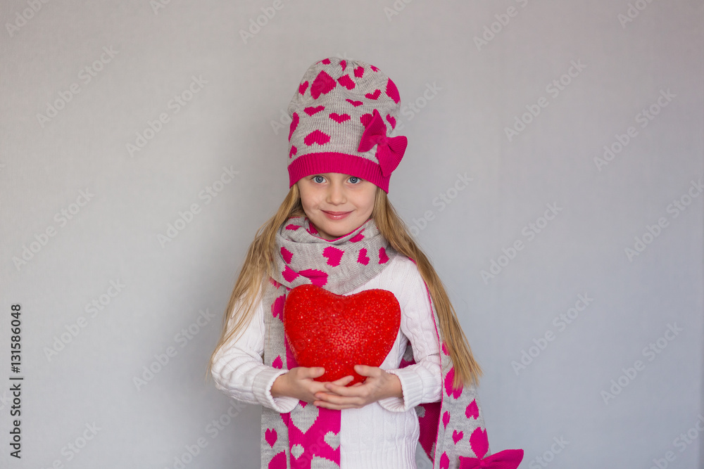 cute little girl in a cap and a scarf with a heart holding a red heart. Valentine's day holiday.