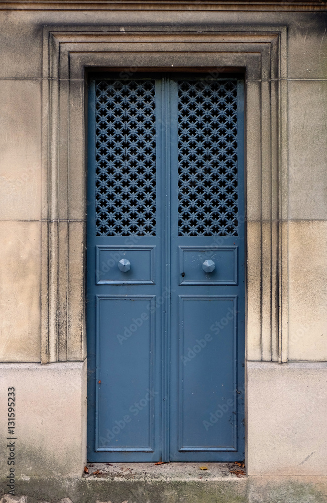 Blue closed iron double door with door knob and hole pattern in a stone wall. The door frame is rectangle and ornated with chiseled lines. The texture is made of small crosses connected by steel bars. Blue closed iron double door with door knob and hole pattern in a stone wall. The door frame is rectangle and ornated with chiseled lines. The texture is made of small crosses connected by steel bars.
